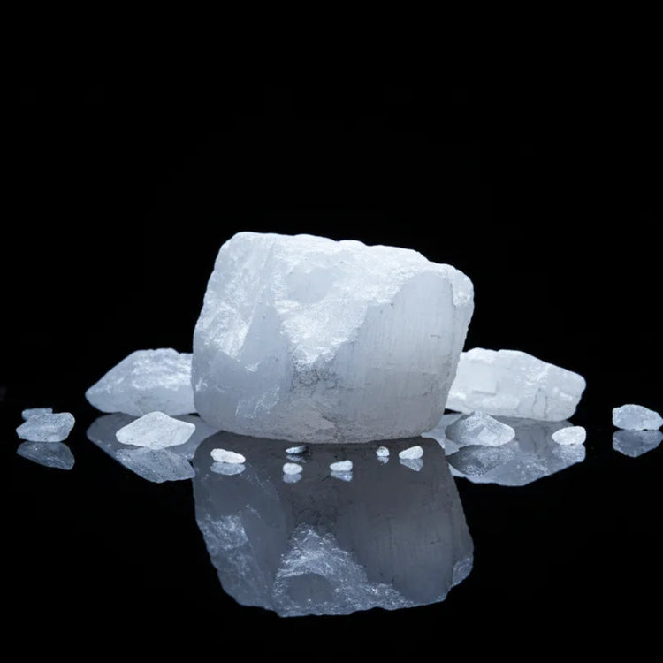 A cluster of rough, translucent white camphor chunks with sharp edges, arranged on a glossy black reflective surface against a solid black background.