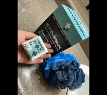 Hand hold a Recovery Shower Steamer next to the product box and blue loofah